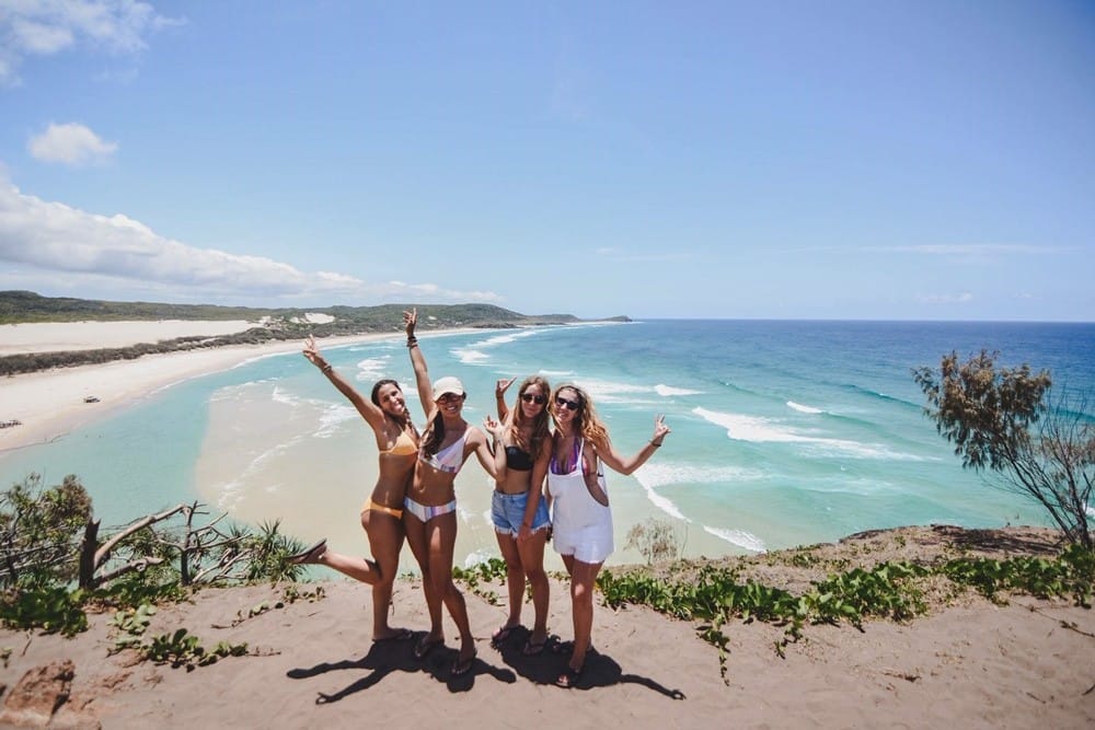 grupo de cuatro chicas en bikini que sonríen a cámara y detrás una playa paradisíaca y el cielo despejado